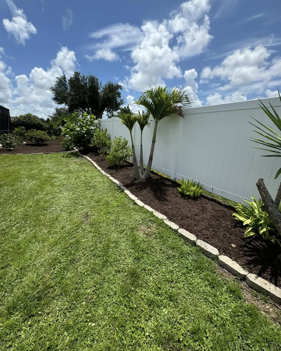 Mulch bed and palms along a white privacy fence